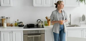 Une femme se tient debout devant sa cuisine. La cuisine blanche est bien rangée.