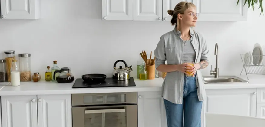 Une femme se tient debout devant sa cuisine. La cuisine blanche est bien rangée.